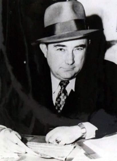 An old B&W photo of a round faced man in a hat, leanign over a desk - Captain Jack Donahoe.