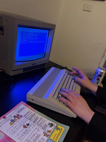 Hands with painted nails on a C64 C model, a Sony PVM as its display. An Usborne BASIC programming book sits open to the left, and a can of beer to the right.