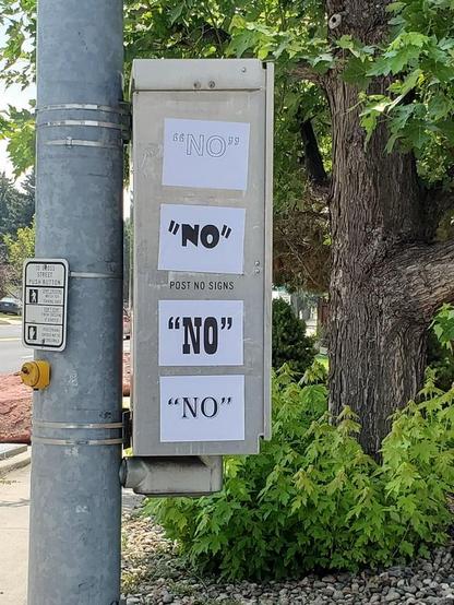A photo of a metal utility box mounted on a pole with four separate sheets of paper taped to it, each reading "NO" in quotation marks, placed directly above and below a stamped metal sign that reads "POST NO SIGNS".